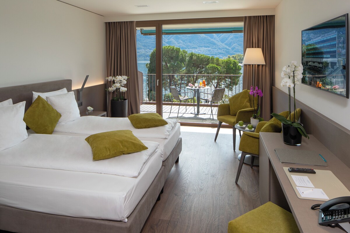 Habitación de hotel con cama, escritorio, silla, televisión y balcón con vistas.