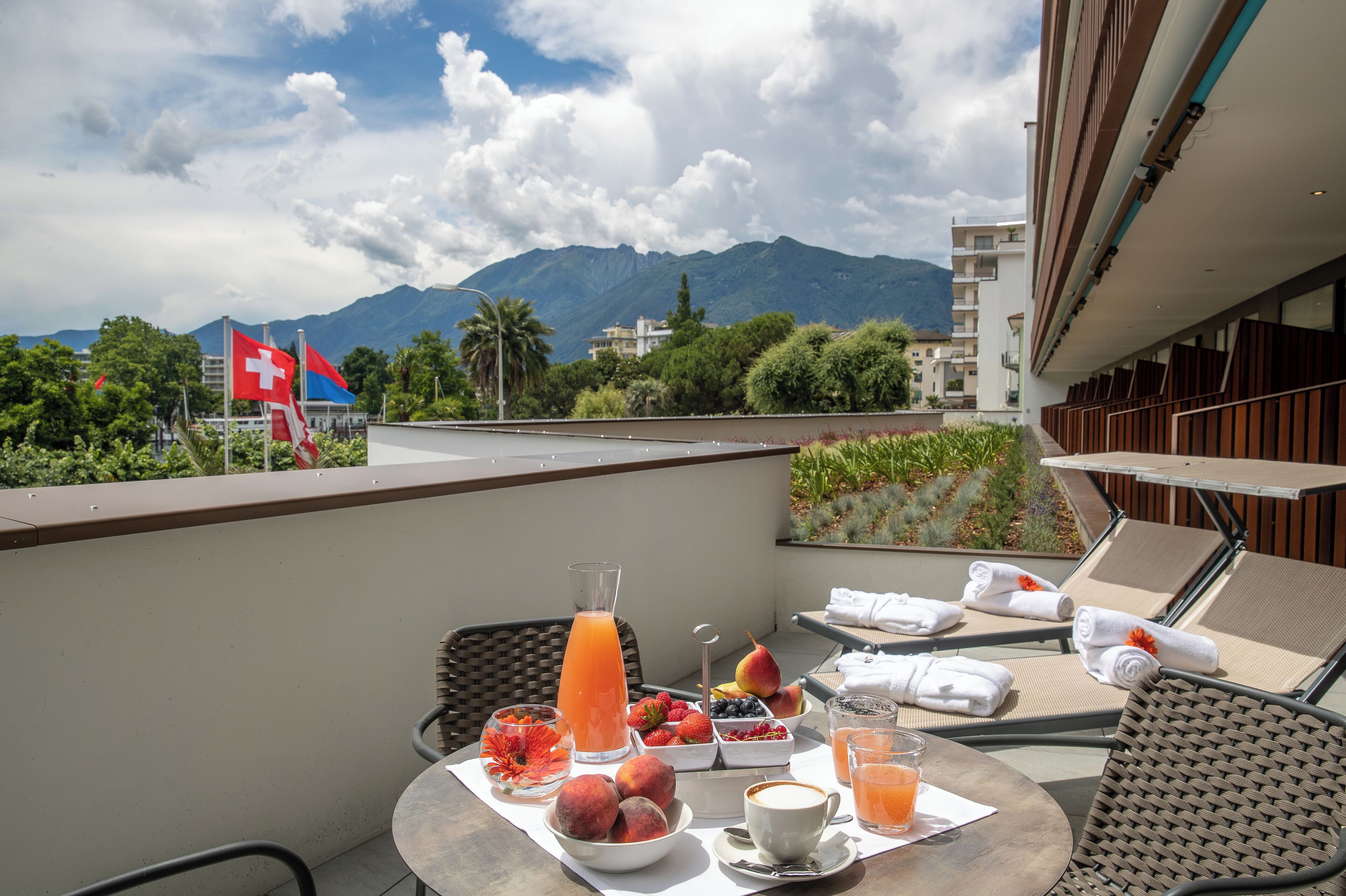 A balcony with a table set for breakfast, including fruits, a coffee cup, and a glass of orange juice. There are lounge chairs and a Swiss flag in the background.