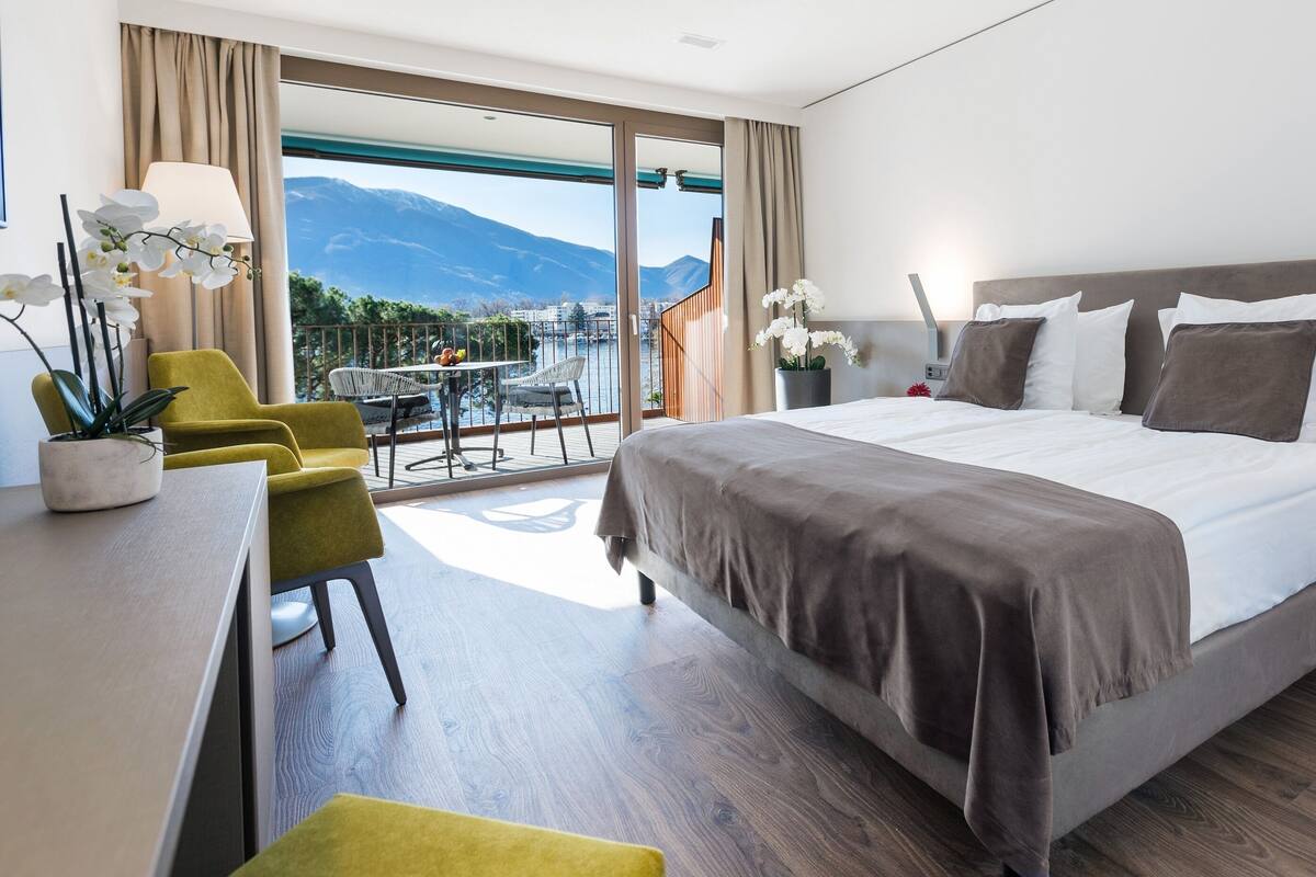 A modern hotel room with a large bed, a desk with a chair, a balcony with a view of mountains, and a dining area with a table and chairs.