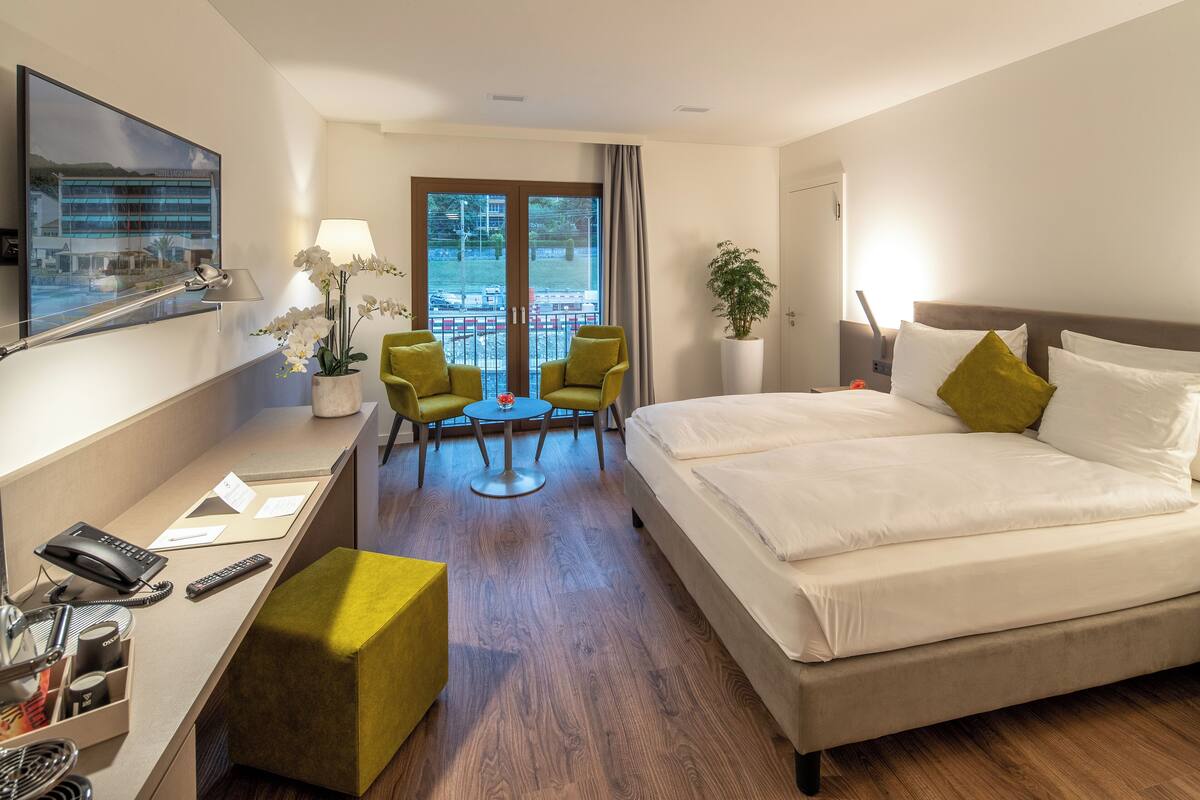 A hotel room with a bed, a desk with a phone and remote, a small table with chairs, a potted plant, and a large window with a view of a building and trees.