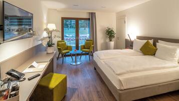Habitación de hotel con cama, escritorio con teléfono y control remoto, pequeña mesa con sillas, planta en maceta y ventana grande con vista a un edificio y árboles.