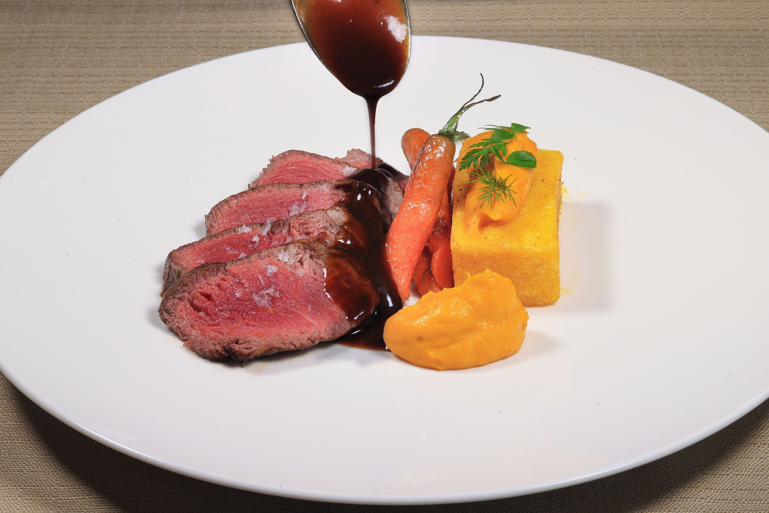 A plate with sliced meat, carrots, and a yellow vegetable, possibly squash, with a sauce being poured over it.