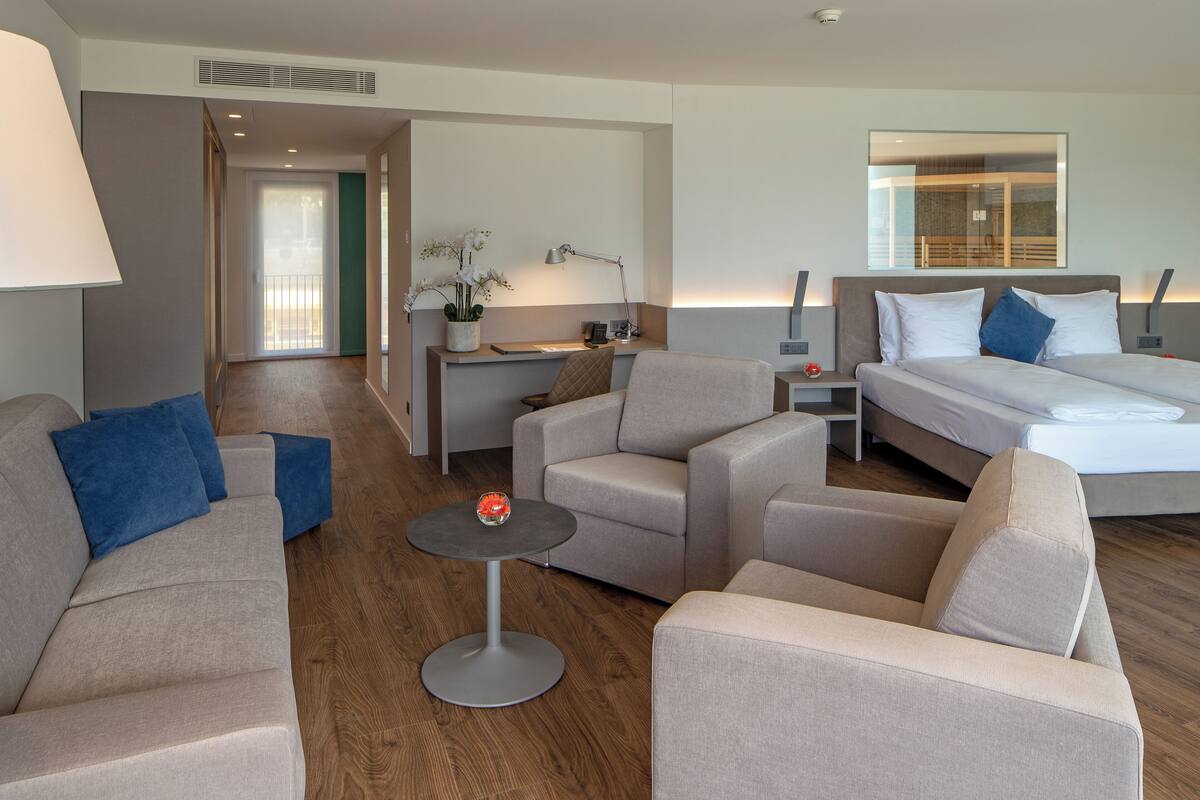A modern hotel room with a sofa, a small round table, a desk with a lamp, a bed, and a large mirror.