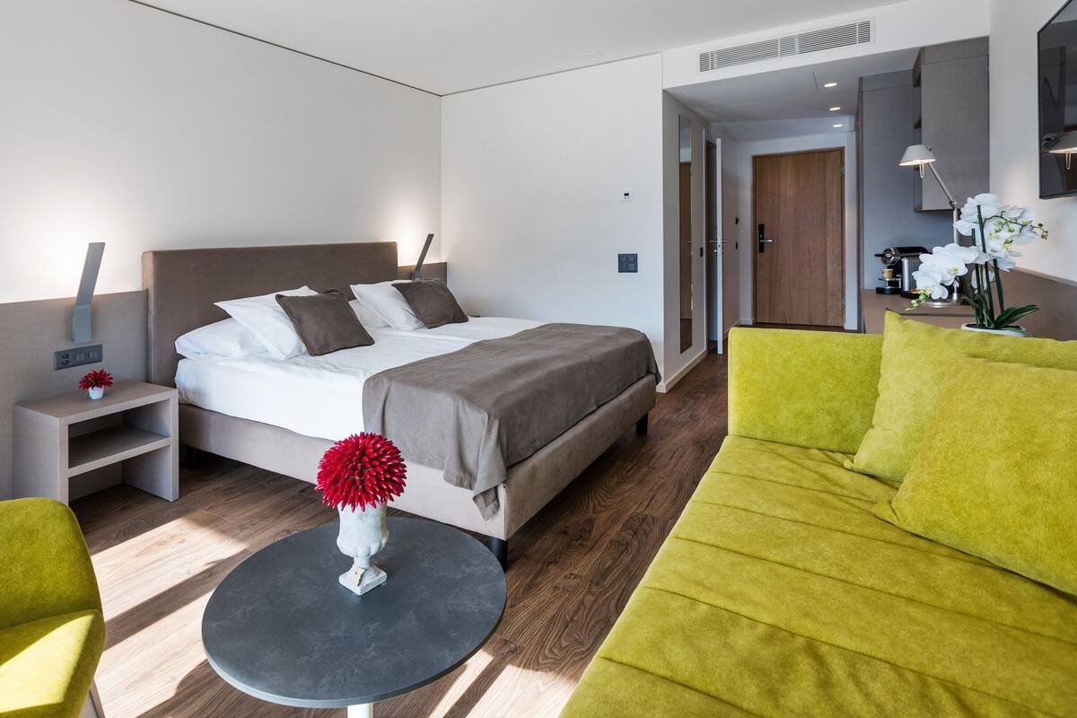 A modern hotel room with a bed, bedside tables, a round table, and a sofa.