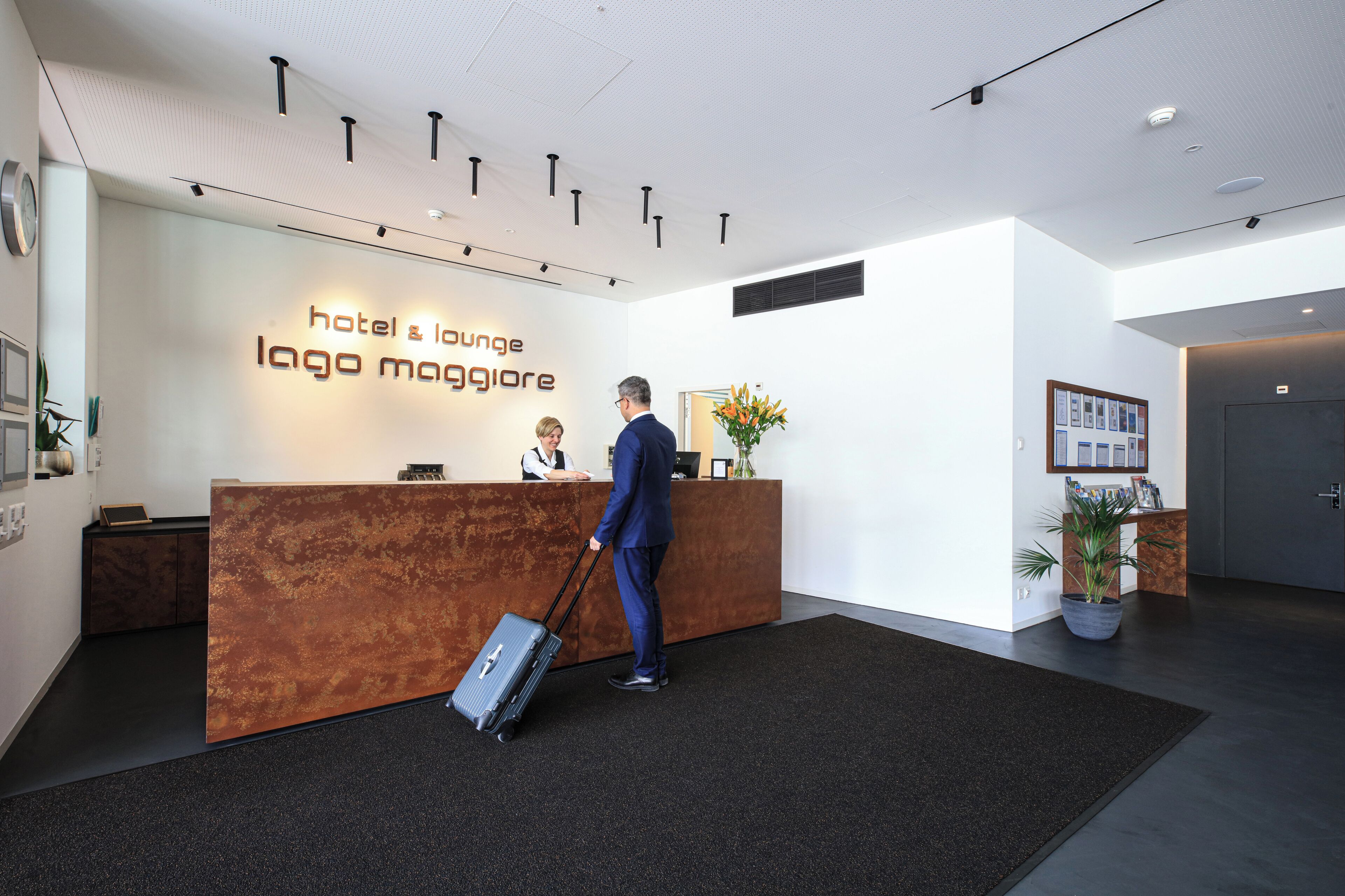 A hotel reception area with a modern design, featuring a reception desk, a man with a suitcase, and a receptionist.