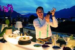A person taking a photo with a smartphone at a table with a dessert plate, a bowl of fruit, and a potted plant.