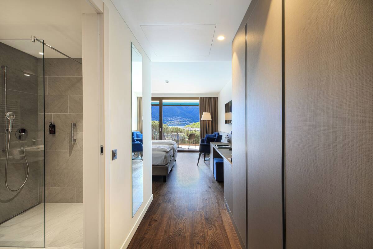 A modern hotel room with a shower, a bed, a desk, and a view of the city.