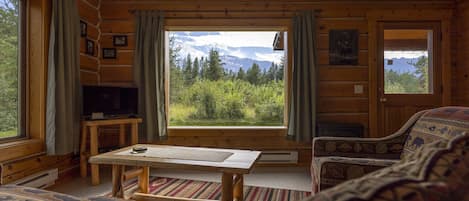 A cozy cabin interior with a large window offering a mountain view, a television, and a rustic wooden table.