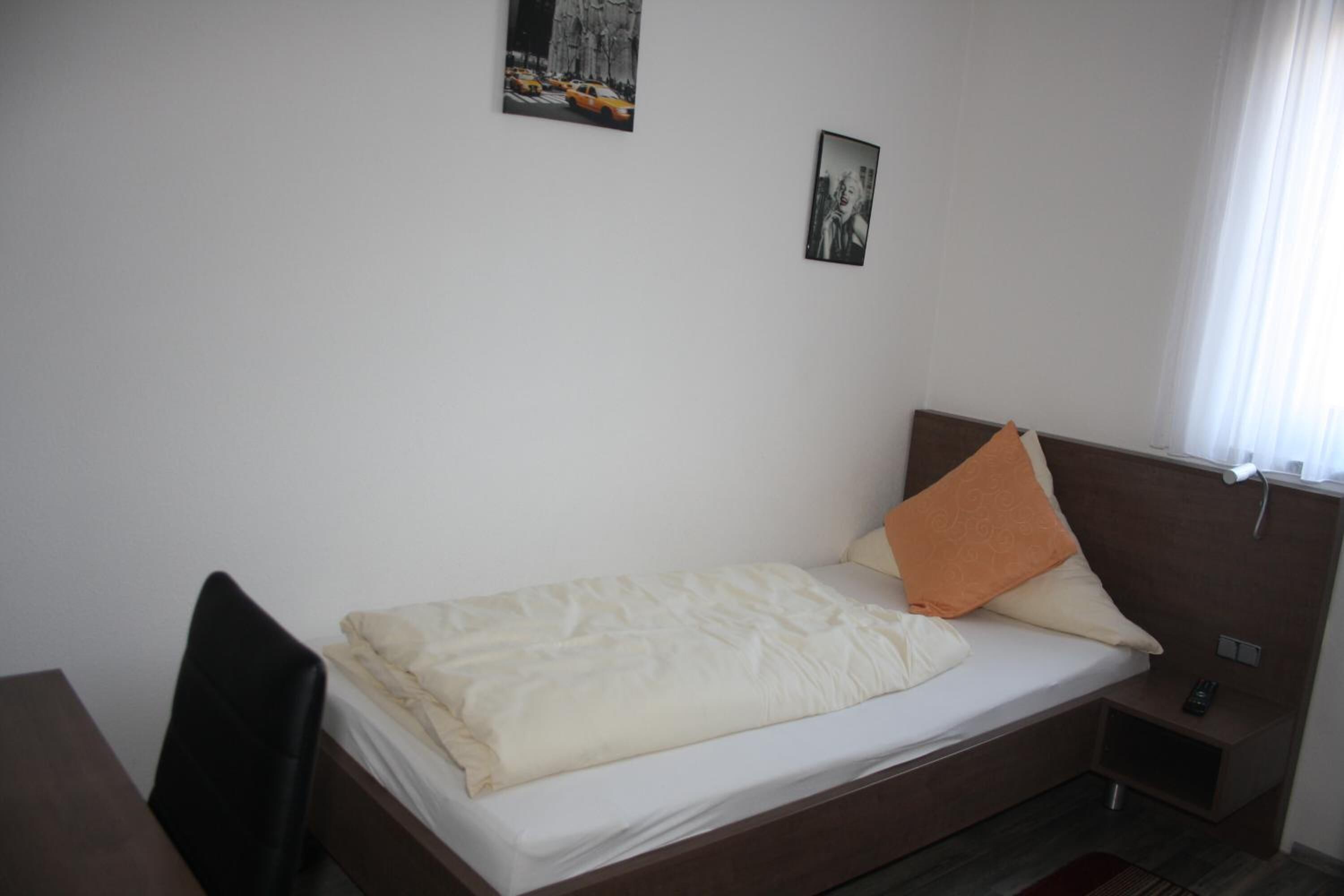 A single bed with a white comforter and two pillows, a small bedside table with a remote control, and two framed pictures on the wall.