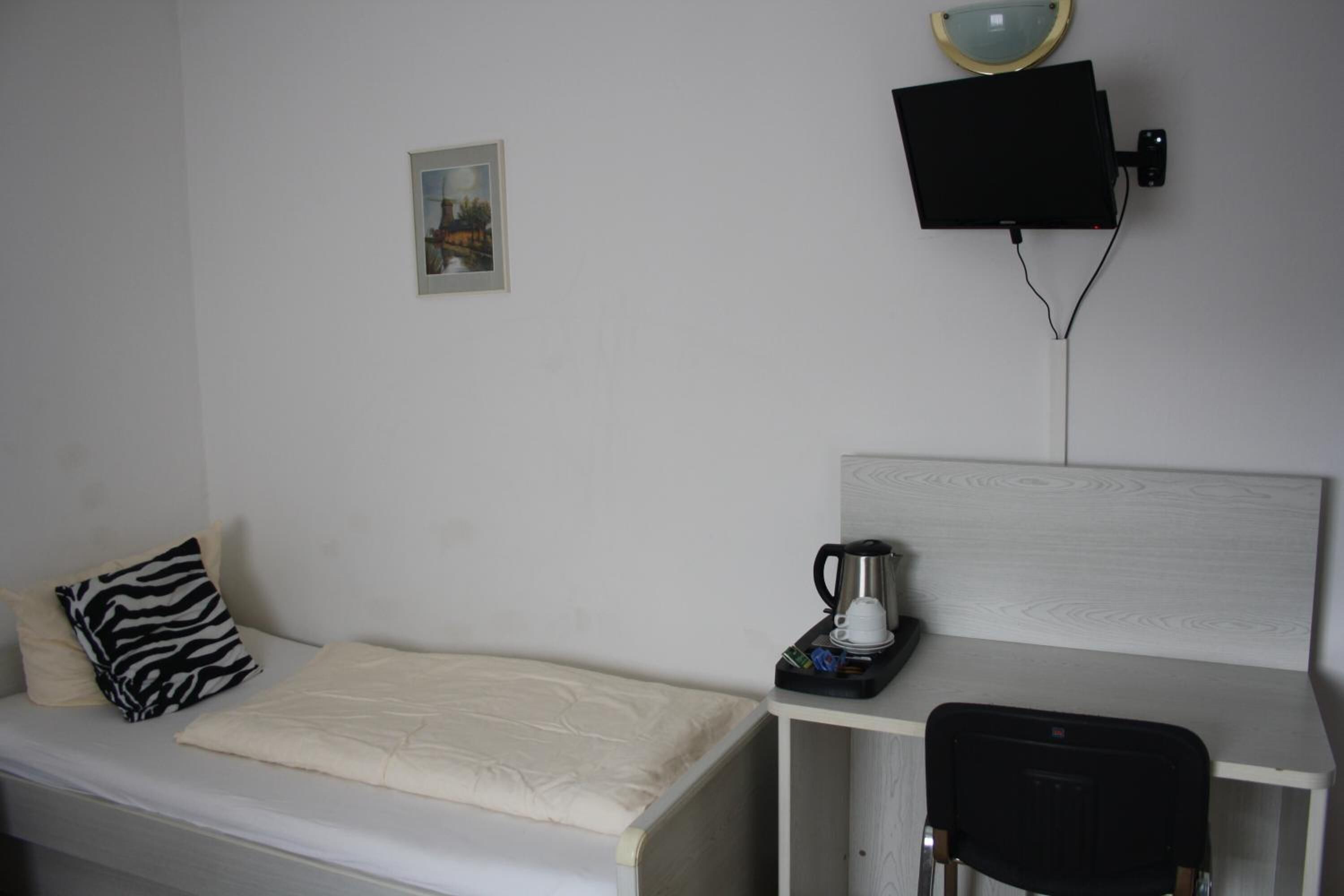 A small, neatly arranged room with a single bed, a bedside table with a coffee maker, a chair, and a mounted television.