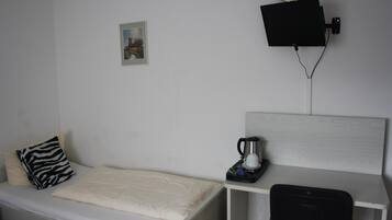Una habitación pequeña y ordenada con una cama individual, una mesita de noche con cafetera, una silla y un televisor de pared.