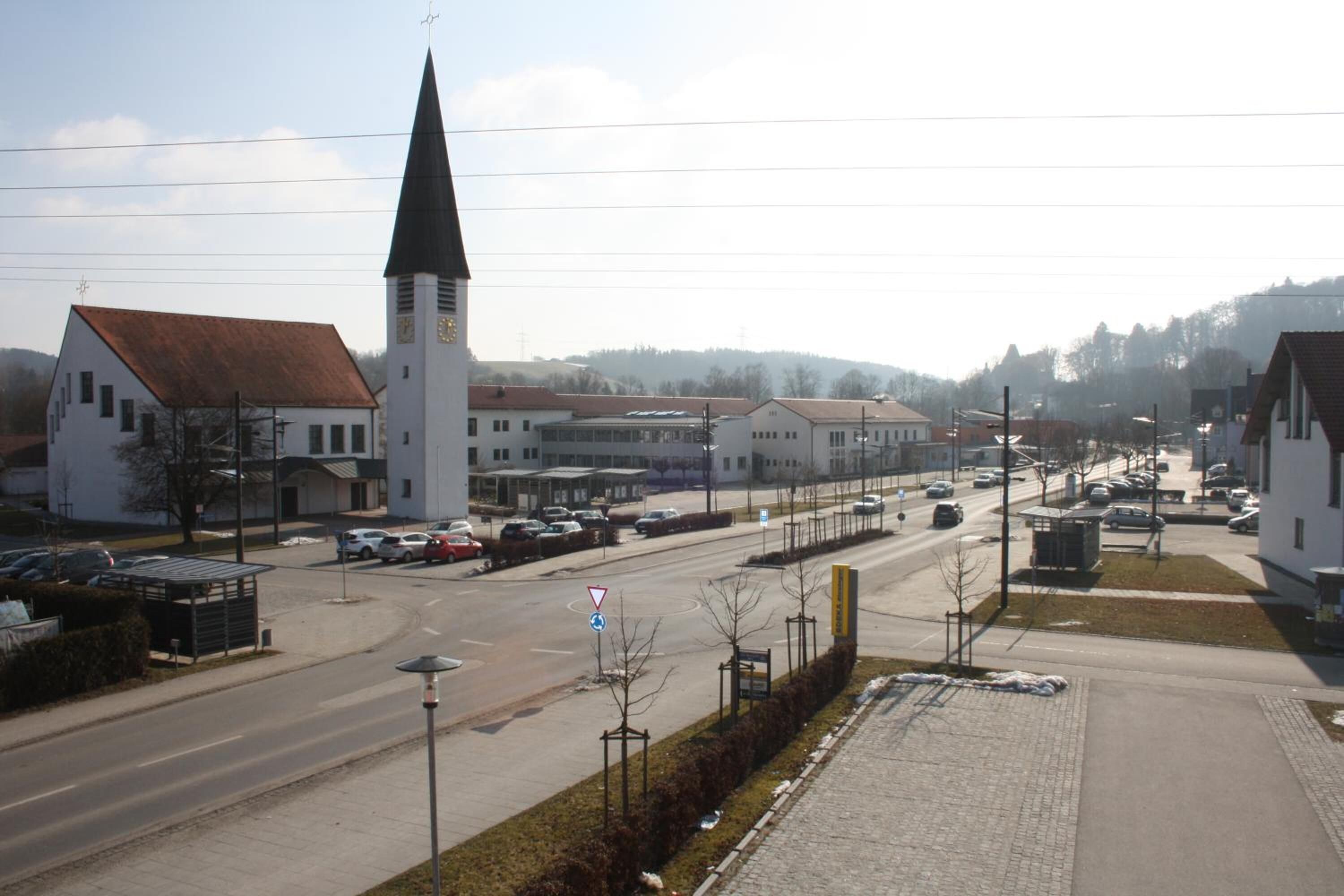 Una plaza de pueblo con una iglesia, una calle con coches y un sendero para peatones.