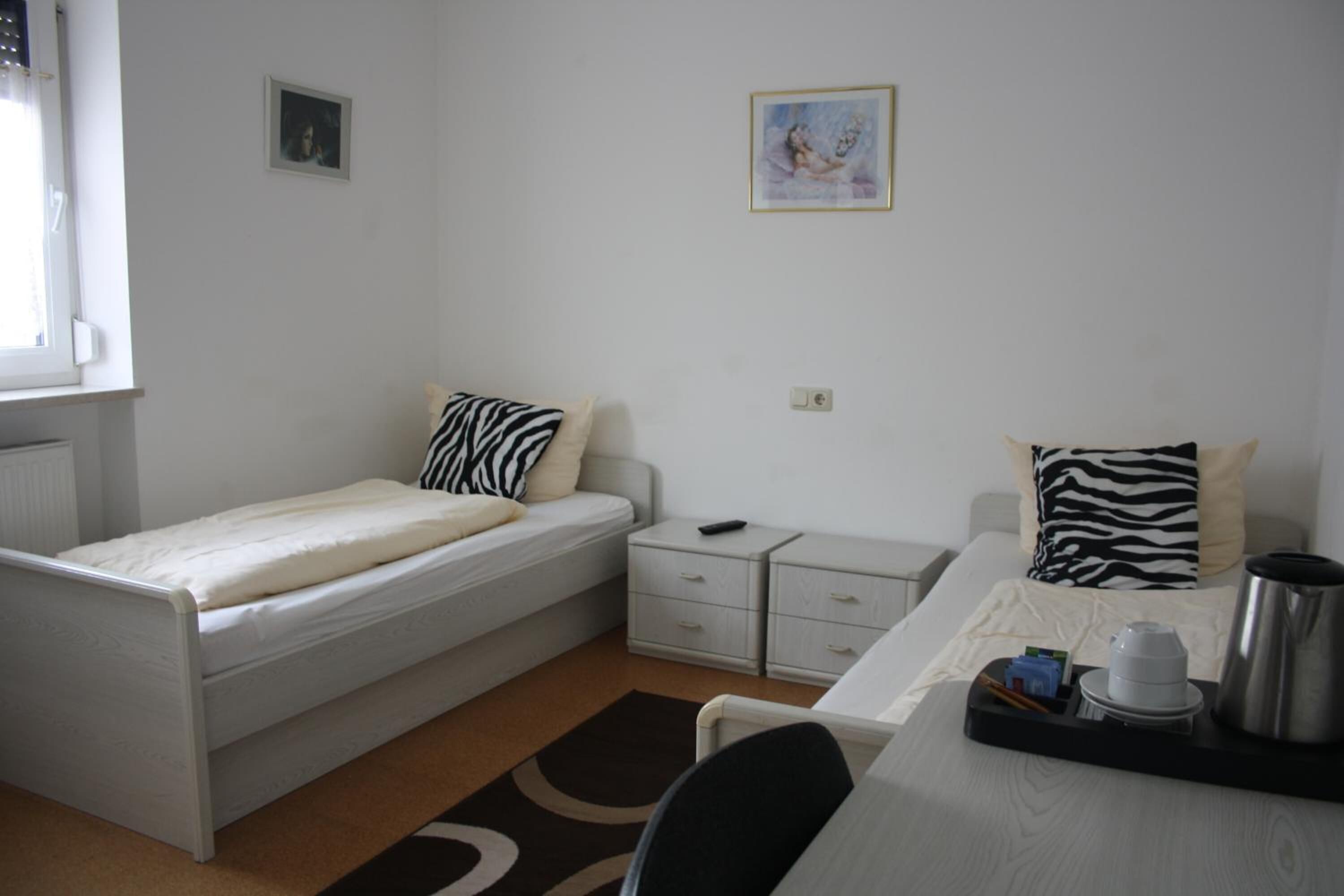 Ein kleines, ordentlich eingerichtetes Schlafzimmer mit einem Einzelbett, einem Nachttisch, einem Stuhl und einem Schreibtisch mit Kaffeekocher und Tassen.