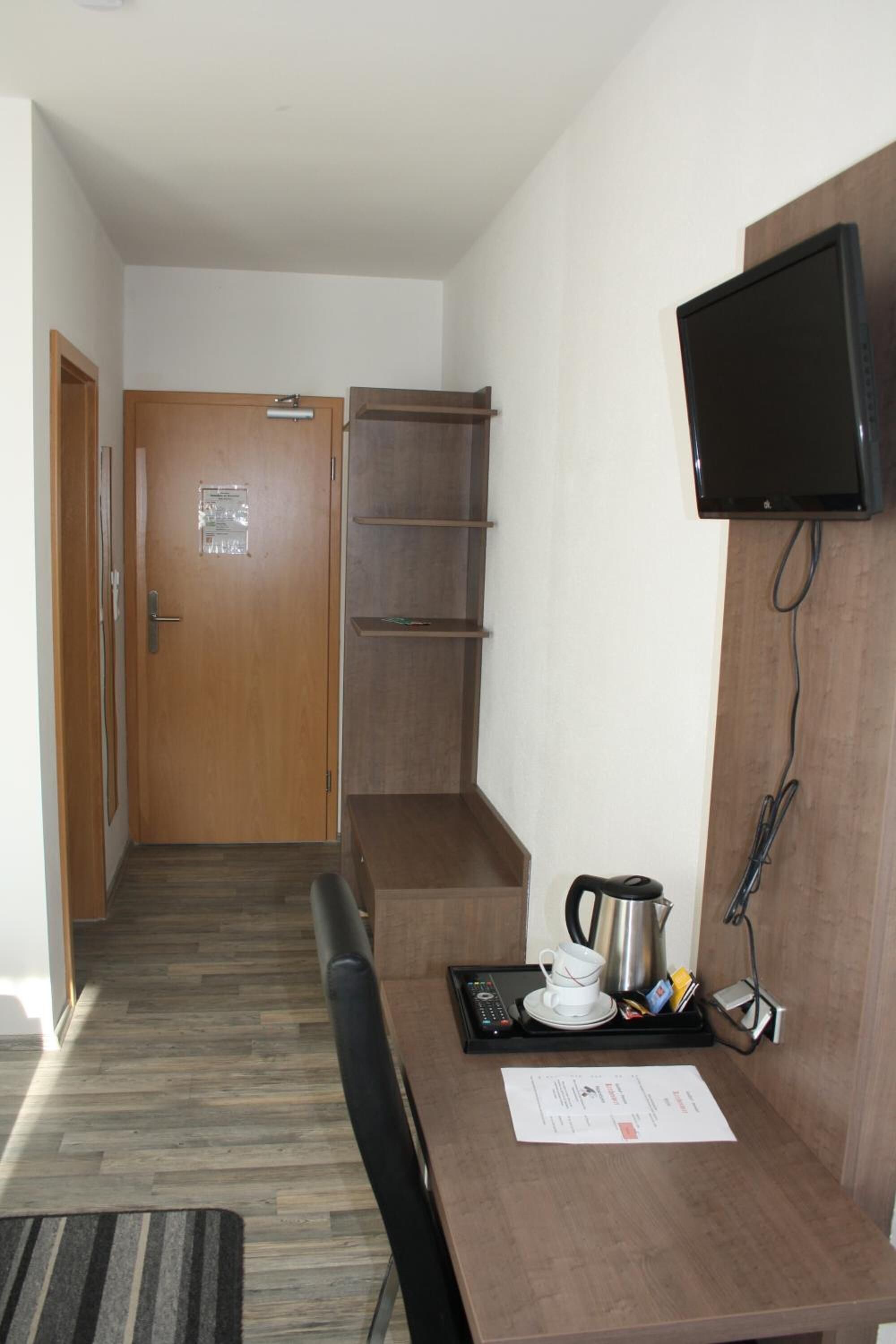 Ein Hotelzimmer mit Holztür, einem an der Wand befestigten Fernseher, einem kleinen Tisch mit Tablett, einer Tasse und einem Wasserkocher.