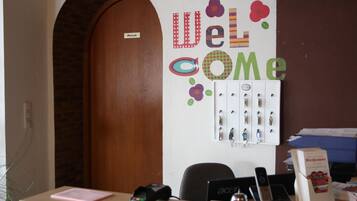 Un mostrador de recepción con un cartel que dice 'Welcome', una puerta privada, un expositor de llaves y una barra con un teléfono, un lector de tarjetas y un libro.