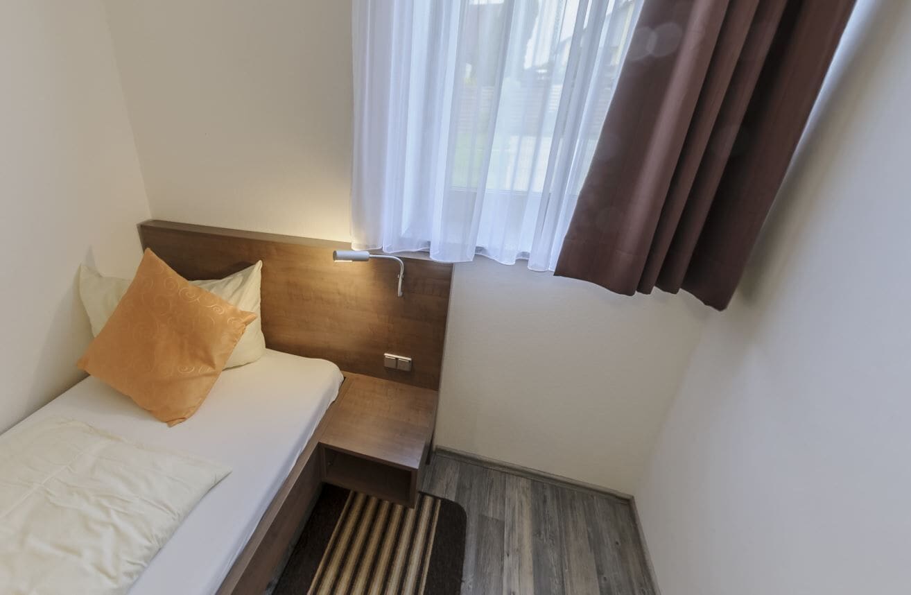 A small, modern hotel room with a single bed, a nightstand, a lamp, and a window with curtains.