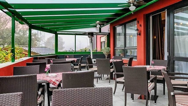 An outdoor patio with wicker furniture and tables, covered by a green roof, and large windows with curtains. - Gasthof-Pension Kirchenwirt (Niederaichbach)