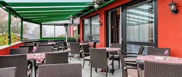 Um pátio externo com móveis de vime e mesas, coberto por um telhado verde, e grandes janelas com cortinas.