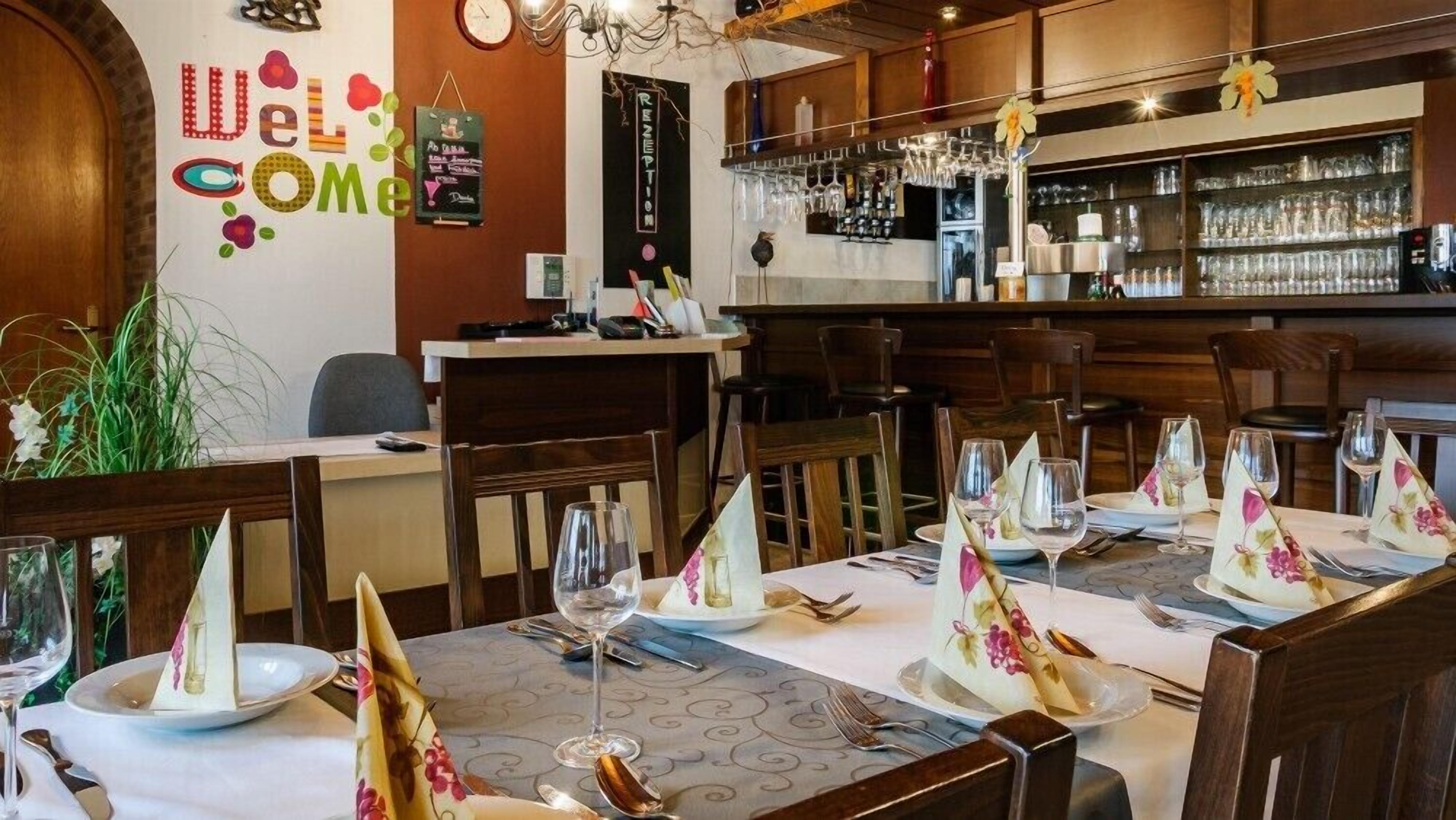 A restaurant with tables set for dining, a bar area with glasses and bottles, and a 'Welcome' sign on the wall.