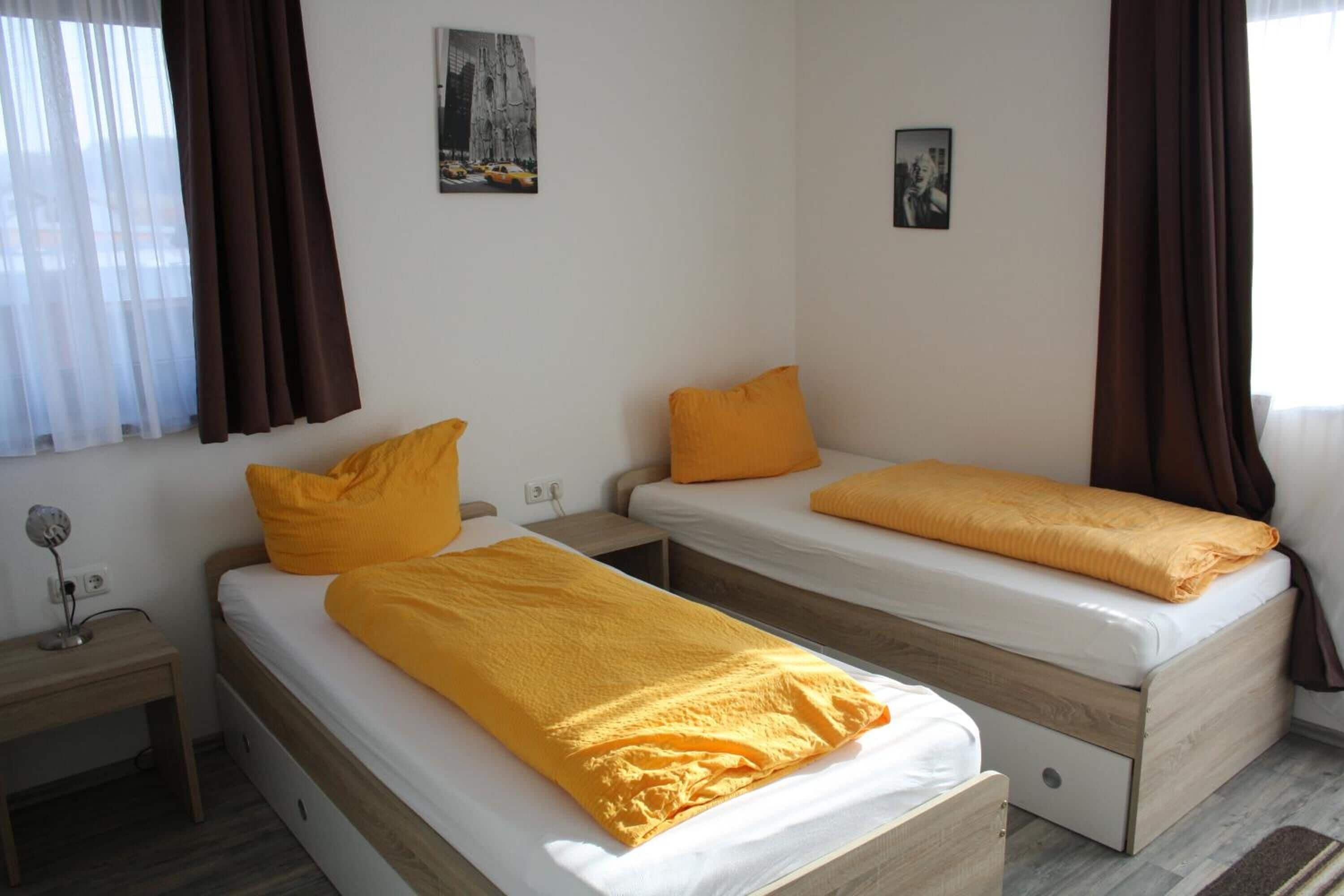 A bedroom with two beds, each with a wooden frame and white bedding. There are two brown curtains, a small table with a lamp, and a wall-mounted picture.
