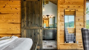 Interior de una cabaña de madera con una cama, un sillón de cuadros, un espejo y vistas a una cocina y montañas.
