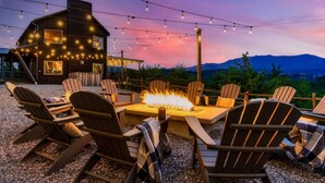 Un espacio al aire libre acogedor con sillas Adirondack y una chimenea, con montañas y una cabaña de madera al fondo.