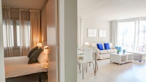 A modern hotel room with a bed, a sitting area with a sofa and chairs, a dining table, and a large window with curtains. - La Gioia 1 by Hello Homes Sitges (Sitges)