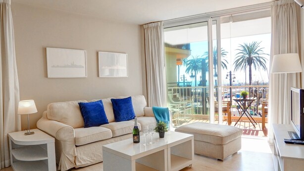 A modern living room with a sofa, coffee table, and TV stand. There is a balcony with a view of palm trees and a pool. - La Gioia 1 by Hello Homes Sitges (Sitges)