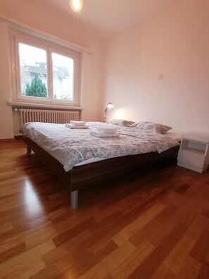 A bedroom with a wooden floor, a bed with a floral bedspread, a bedside lamp, and a window with a view of greenery. - House at Rotsee Lake Lucerne (Lucerne)