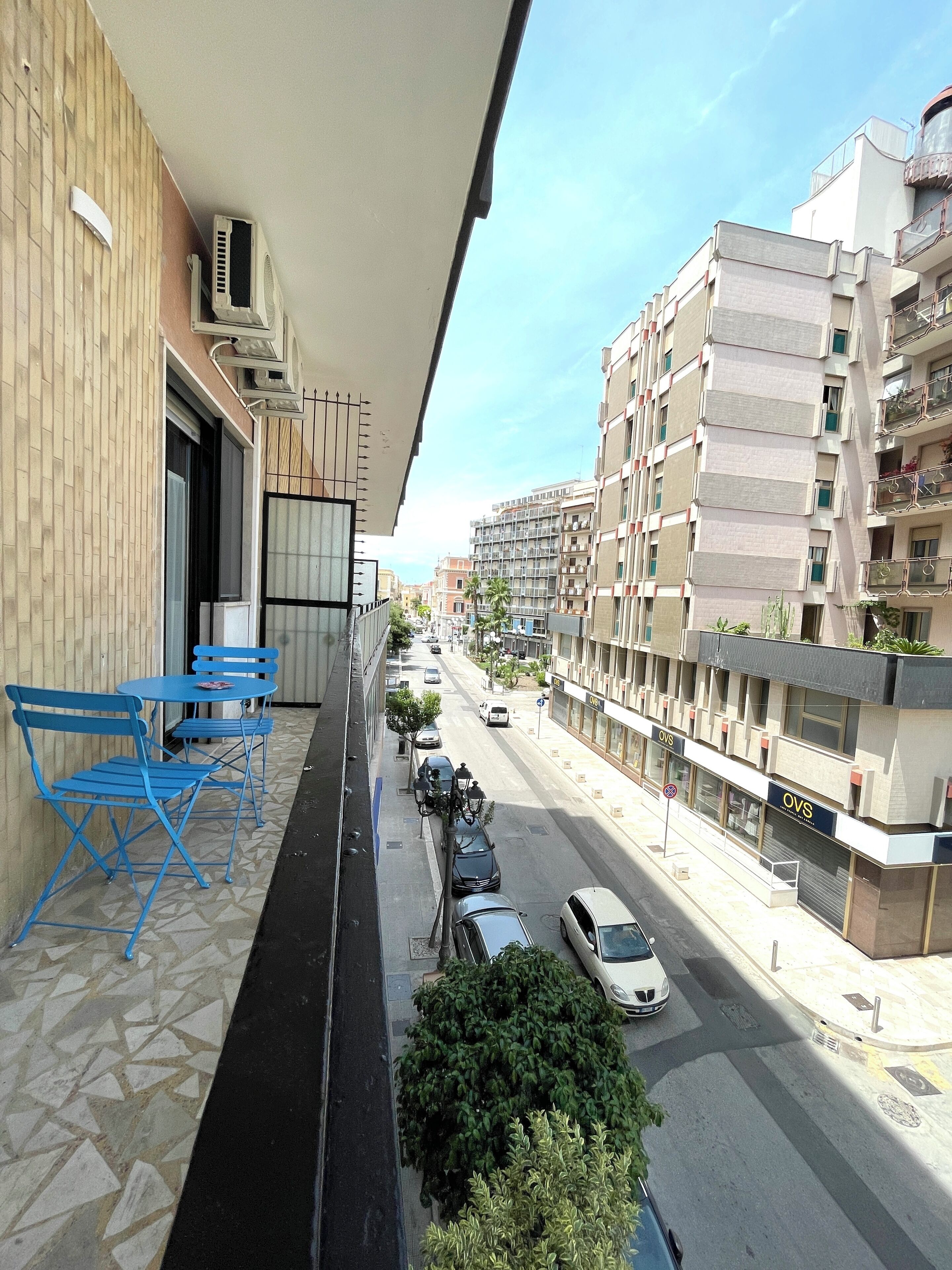 Un balcón con sillas azules, piso de baldosas y vista a una calle con coches estacionados y edificios.