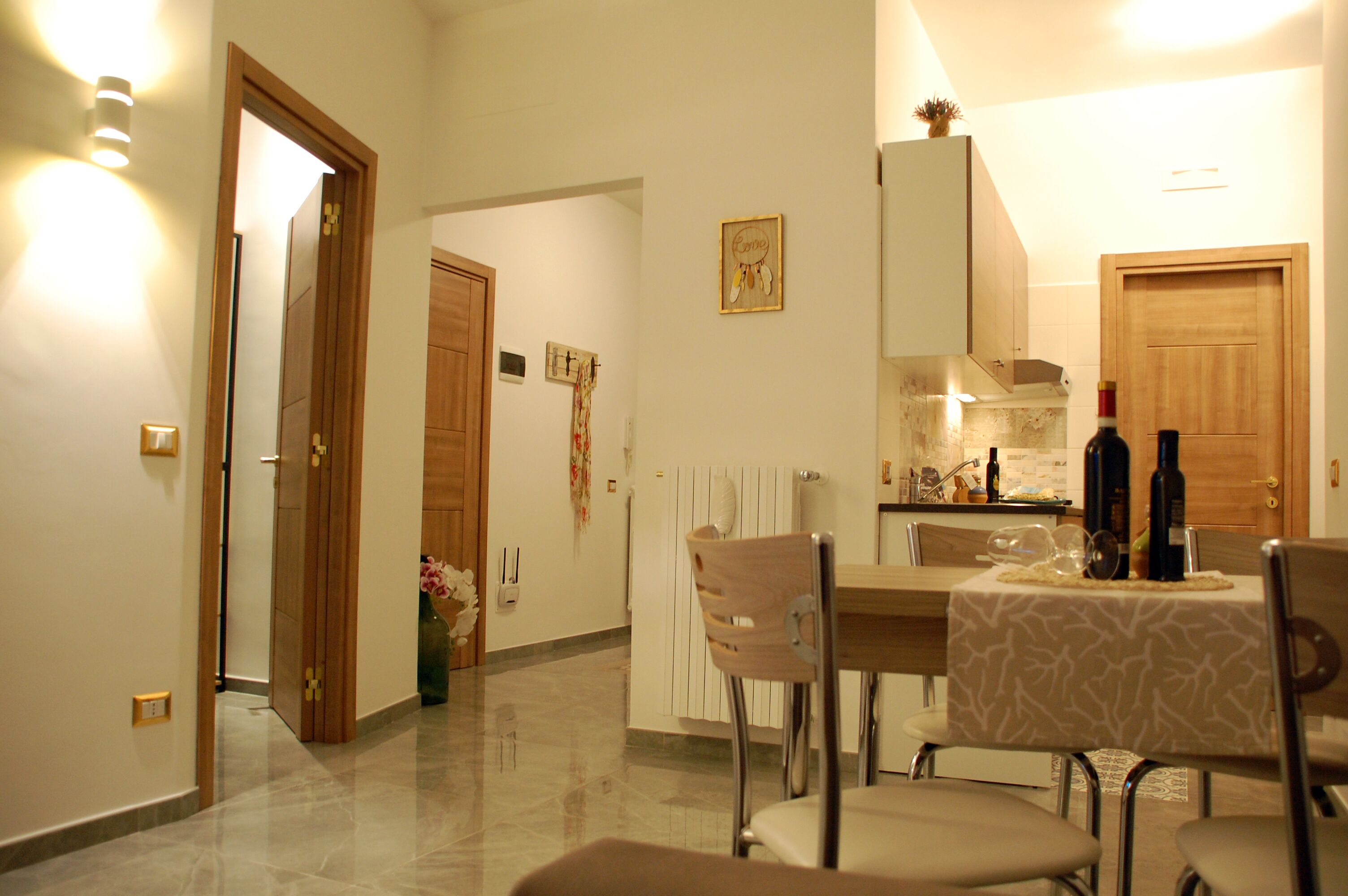 A dining area with a table set for four, a kitchen in the background, and a wooden door leading to another room.