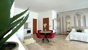 A modern living room with a beige sofa, a dining table with red chairs, a large green plant, and a bed with white bedding. - Dimore del Sud (Trani)