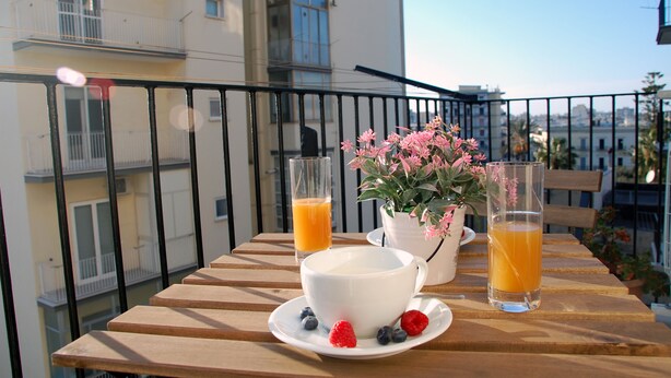 A balcony with a wooden table set for breakfast, including a white cup, two glasses of orange juice, and fresh berries. - Dimore del Sud (Trani)