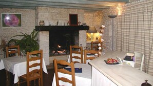 A rustic dining area with a stone fireplace, wooden chairs, a table set for two, and a bar area with a lamp and decorative items. - Le Chambellan (Coux-et-Bigaroque-Mouzens)