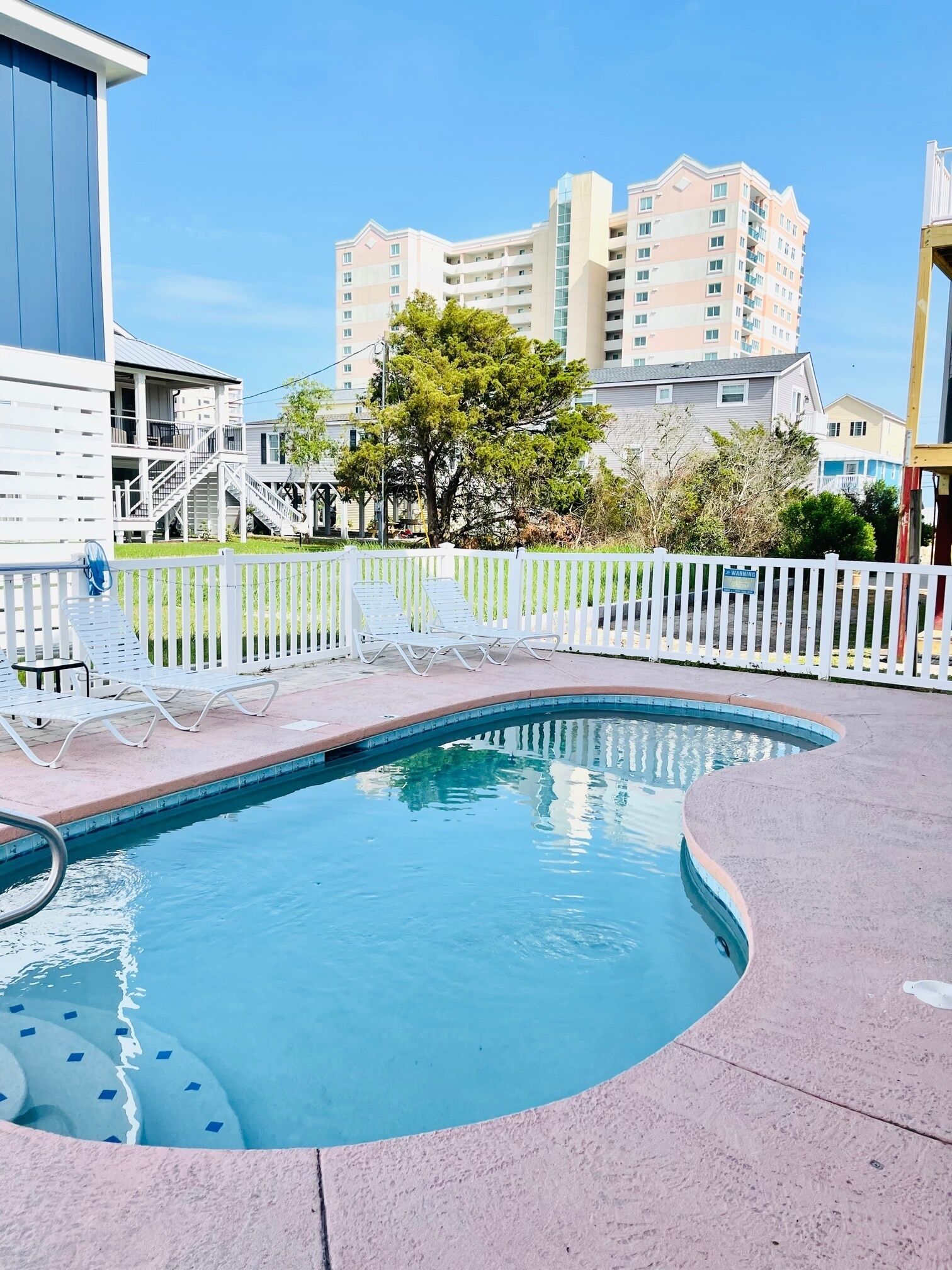 A swimming pool with lounge chairs, a white fence, and a building with multiple floors in the background.