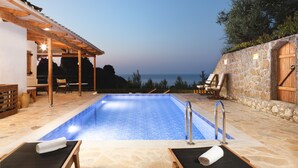 Una piscina con una pérgola y sillones de descanso, rodeada de muros de piedra y vistas al mar.