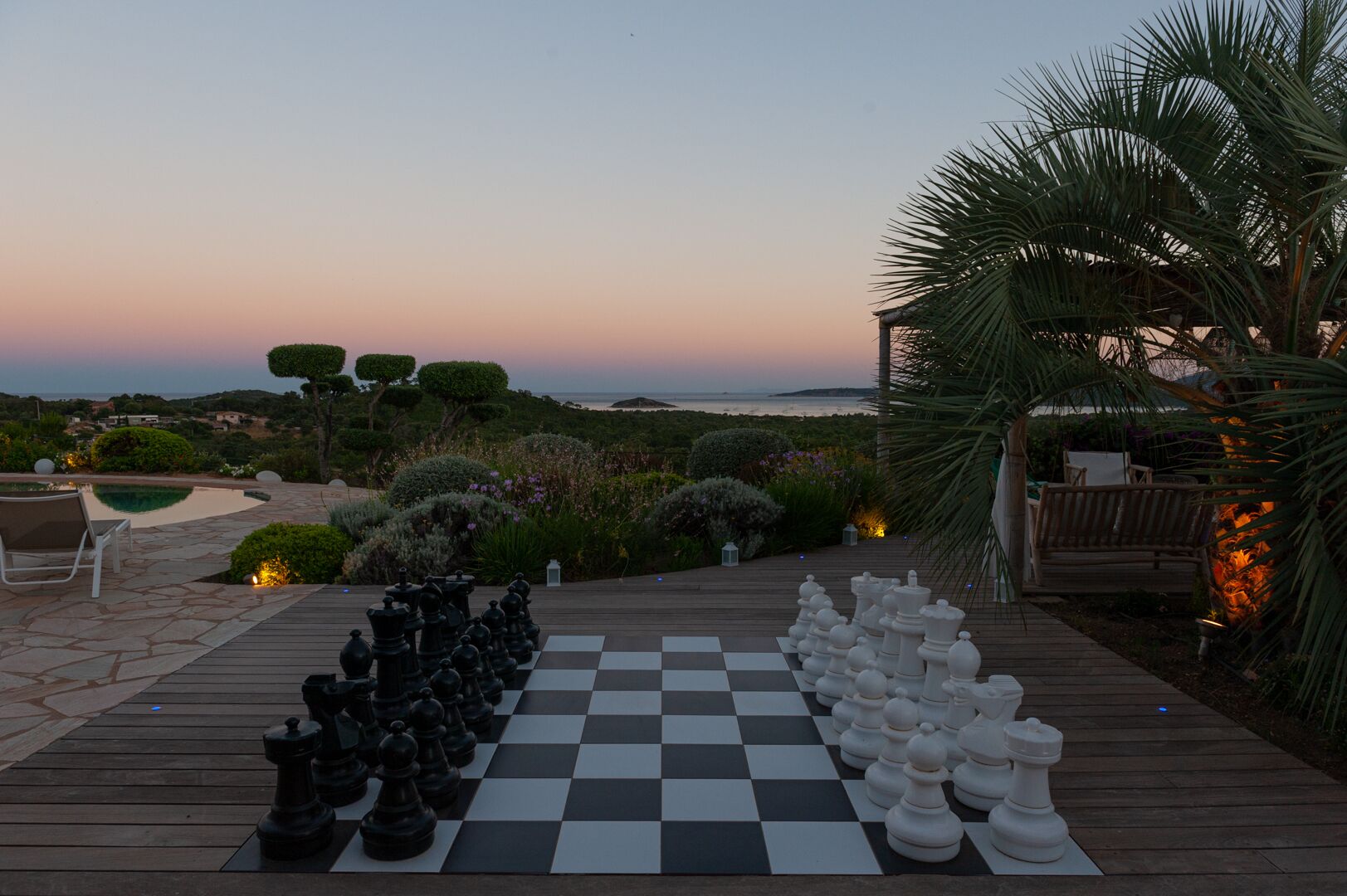 Um jogo de xadrez montado ao ar livre, com uma vista panorâmica do oceano e colinas ao fundo.