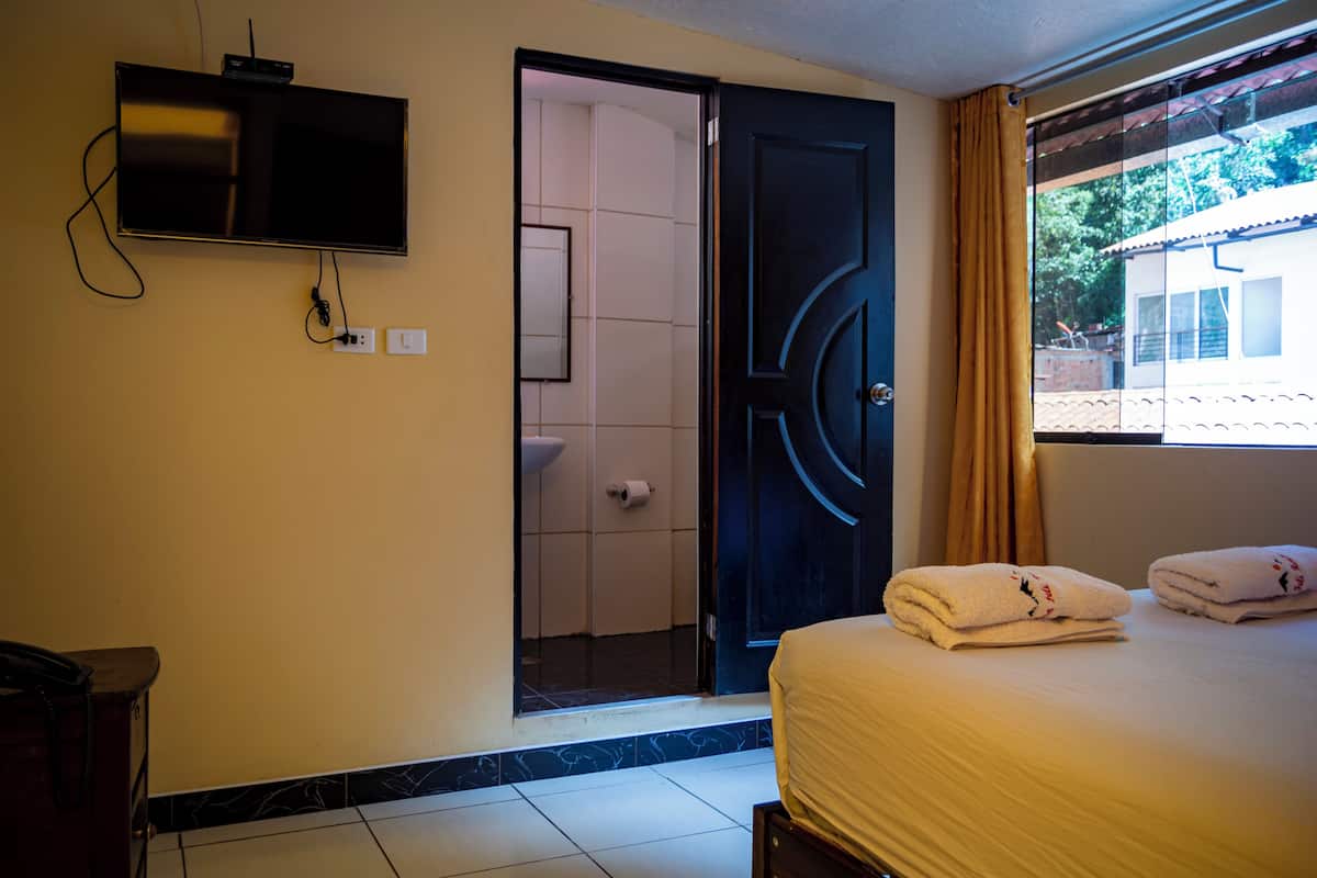 Habitación de hotel con cama, televisión montada en la pared, baño con inodoro y lavabo, y ventana con cortinas.