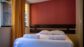Una cama bien hecha con ropa blanca y dos toallas, un cabecero de madera y una pared roja.