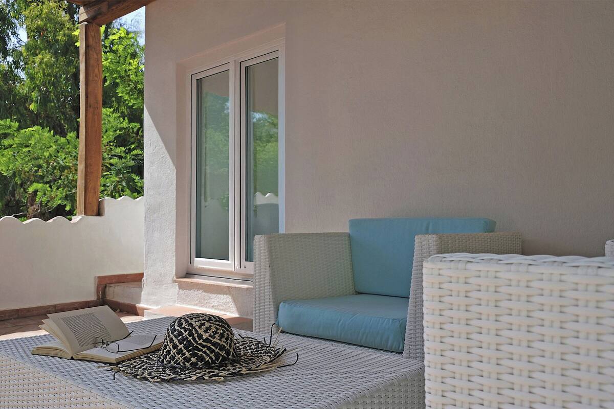 A sunlit patio with a wicker sofa, a reading area with a book and glasses, and a view of greenery through a glass door.
