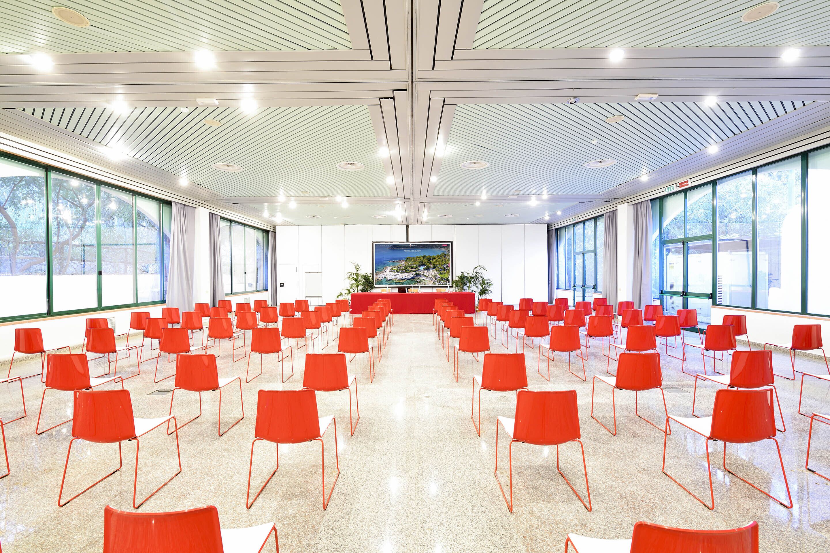 Une salle de conférence avec des rangées de chaises rouges, de grandes fenêtres et un pupitre central.
