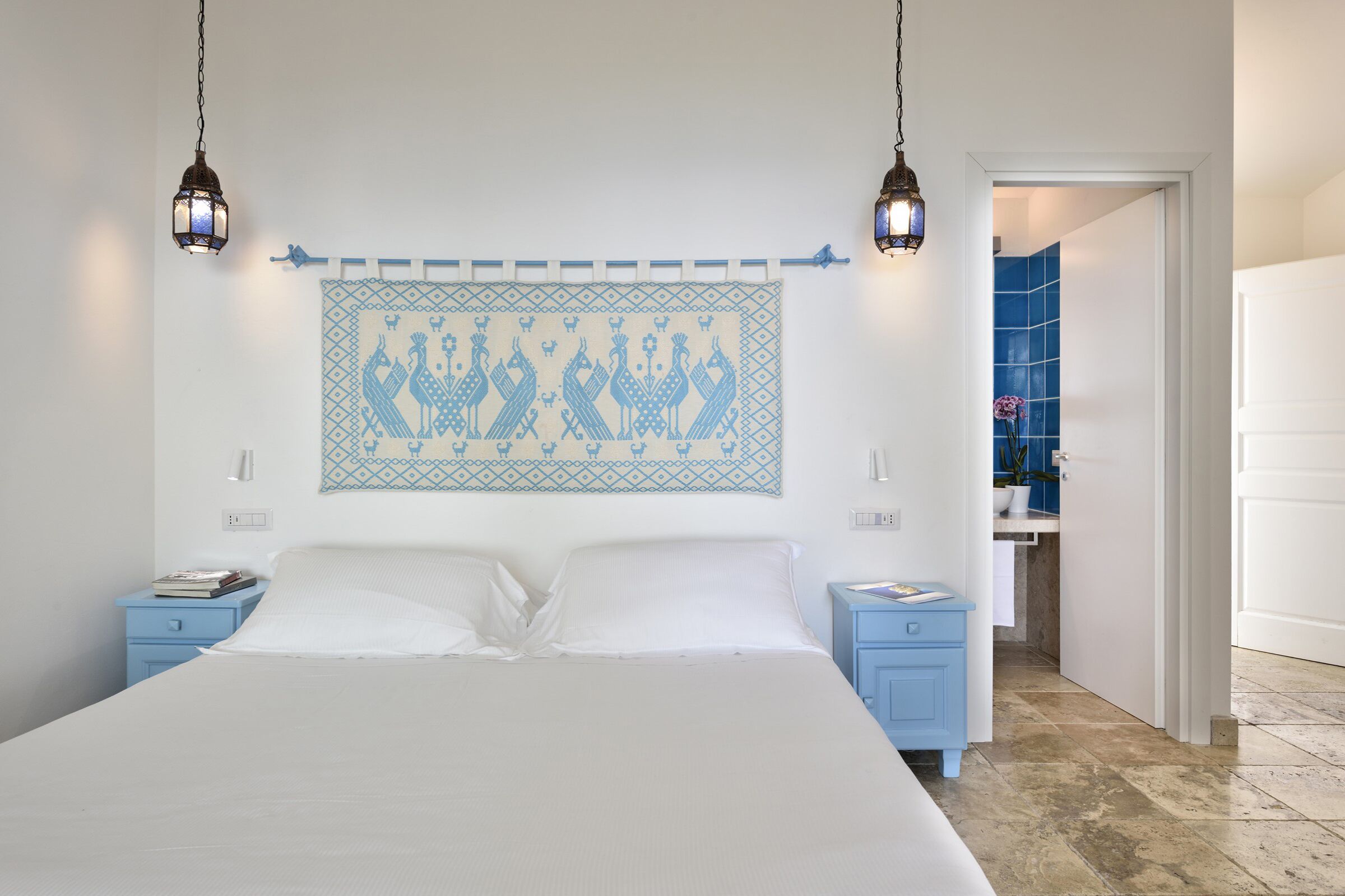 Une chambre à coucher avec un grand lit, deux tables de chevet, une œuvre d’art accrochée au mur et une salle de bain visible à travers une porte ouverte.