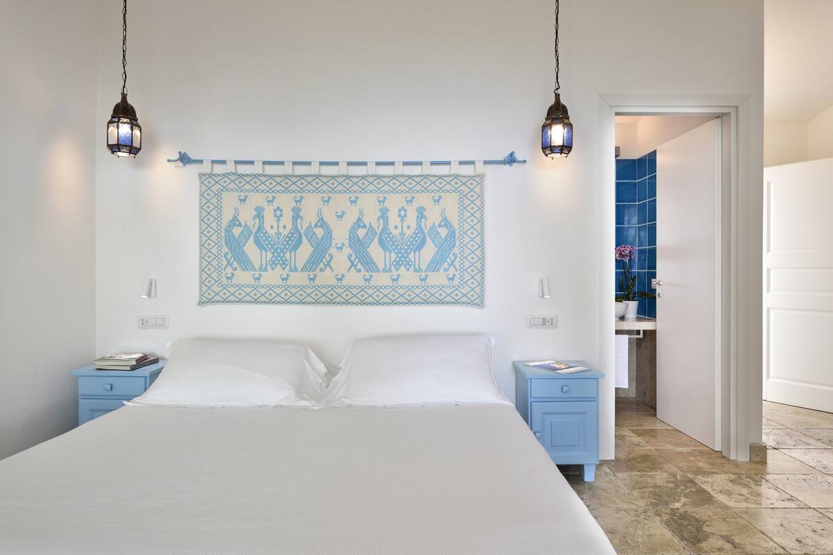 A bedroom with a large bed, two bedside tables, a wall-mounted artwork, and a bathroom visible through an open door.