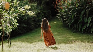 Eine Person in einem langen Kleid geht auf einem Gartenweg, der von viel Grün umgeben ist.