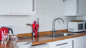 A kitchen with white cabinets, a wooden countertop, a stainless steel sink, a red kettle, a white microwave, and a utensil holder with red utensils. - Pebbles by Bloom Stays - sleeps 2 with the most amazing sea views! (Folkestone)