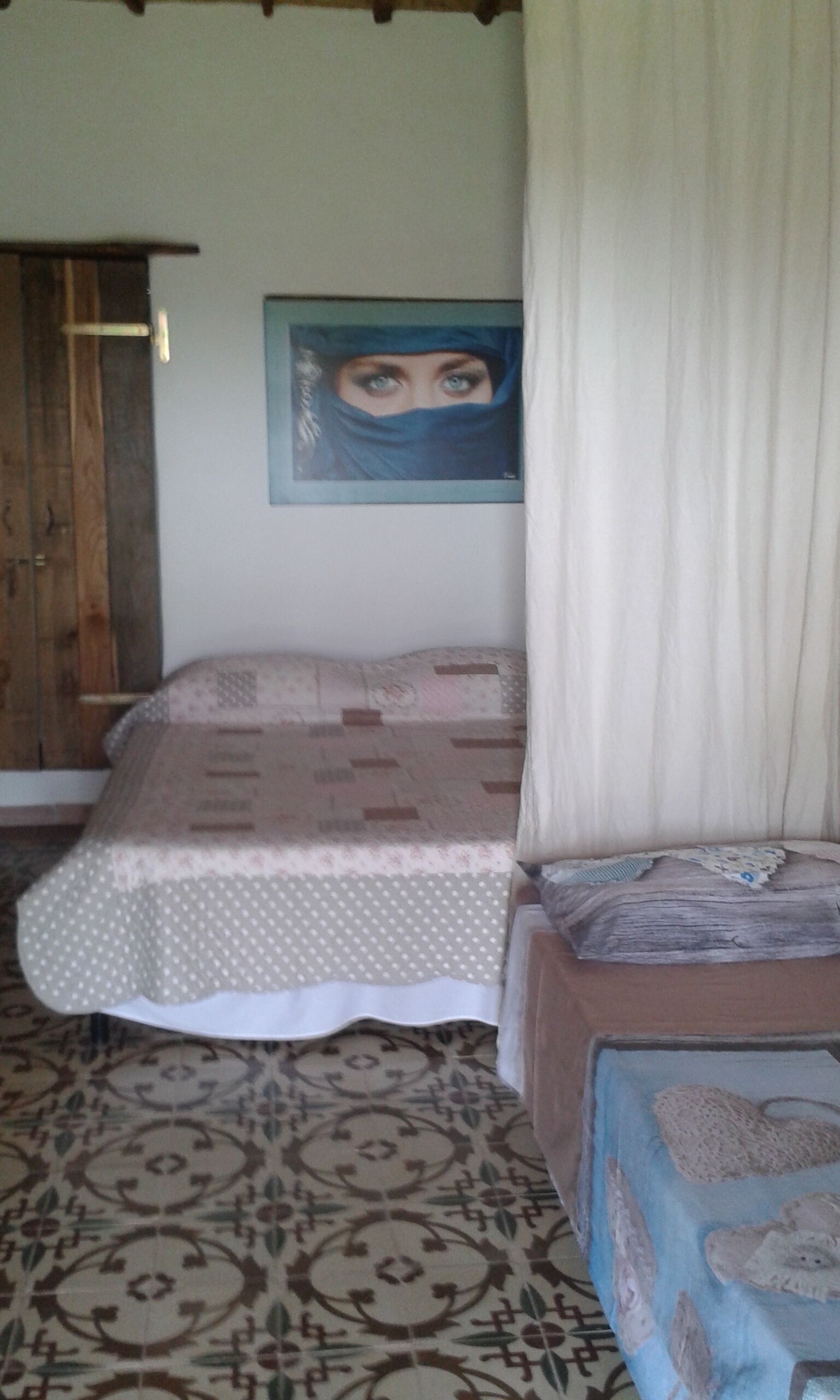 A bedroom with a bed, a wooden wardrobe, a painting on the wall, and patterned floor tiles.