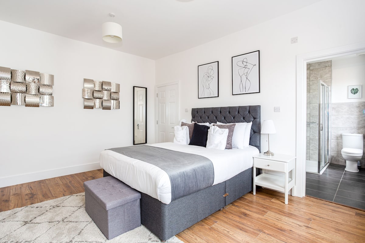 A modern bedroom with a large bed, a bedside table, a bench, and a shower area.