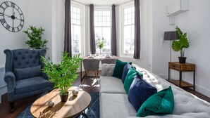 A modern living room with a tufted sofa, a classic armchair, a wooden coffee table, and a dining area with a table set for two. - The Regency Apartments (Southend-on-Sea)