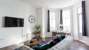 A modern living room with a large flat-screen TV, a round coffee table, a tufted sofa, and a dining area with a table and chairs. - The Regency Apartments (Southend-on-Sea)