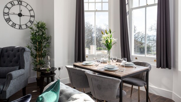 A dining area with a wooden table set for two, a grey sofa, a large wall clock, and a window with curtains. - The Regency Apartments (Southend-on-Sea)